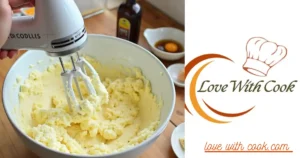 A mixing bowl with creamed butter and sugar, with a hand mixer beside it. A cracked egg and a bottle of vanilla extract are ready to be added